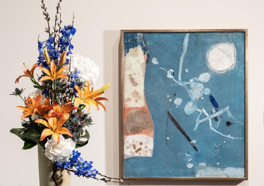 a tall skinny floral arrangement with lillies and other flowers is in front and to the left of an abstract artwork