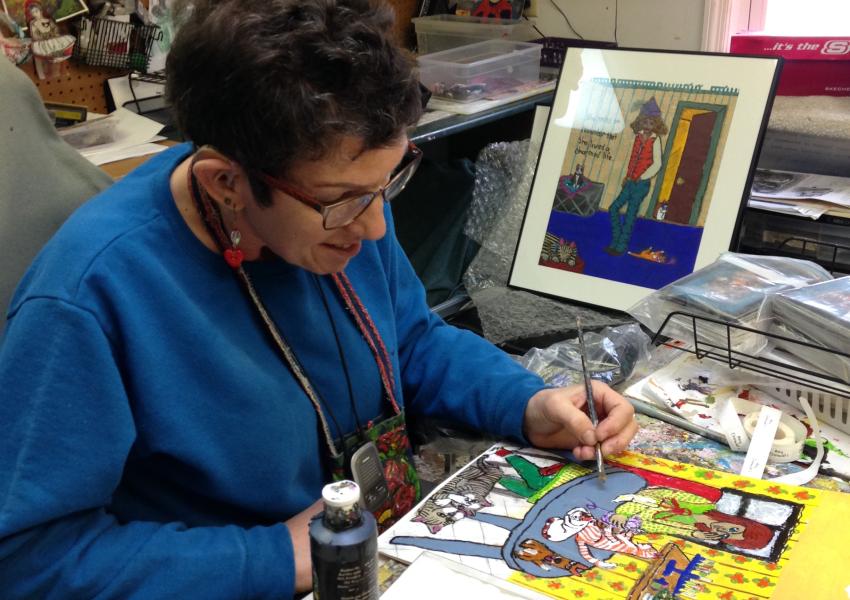 An artist is working in a painting at a desk surrounded by art supplies.