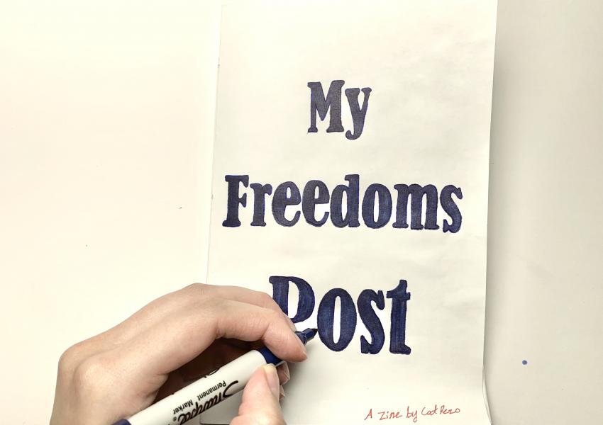 A hand is writing "my freedom post" in sharpie on a set of folded papers.