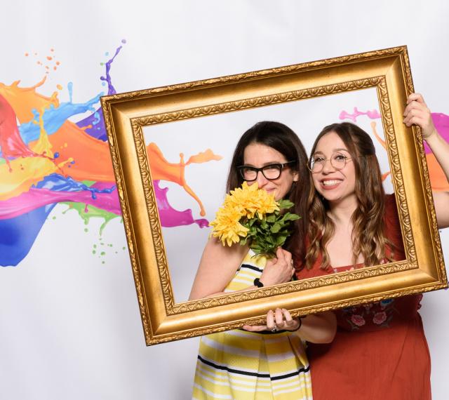Two young women are posing in front of a colorful backdrop with an empty picture frame and fake flowers