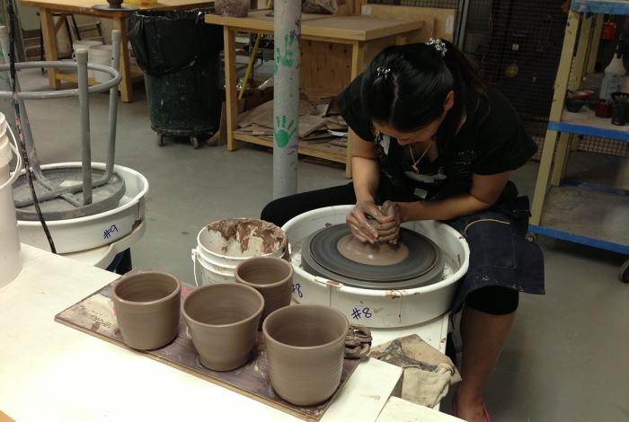 Intermediate Wheel Throwing: View of student throwing clay on the potter's wheel. They sit hunched over, cupping the clay between their hands. 4 previously thrown vessels sit on the bench in front of them.