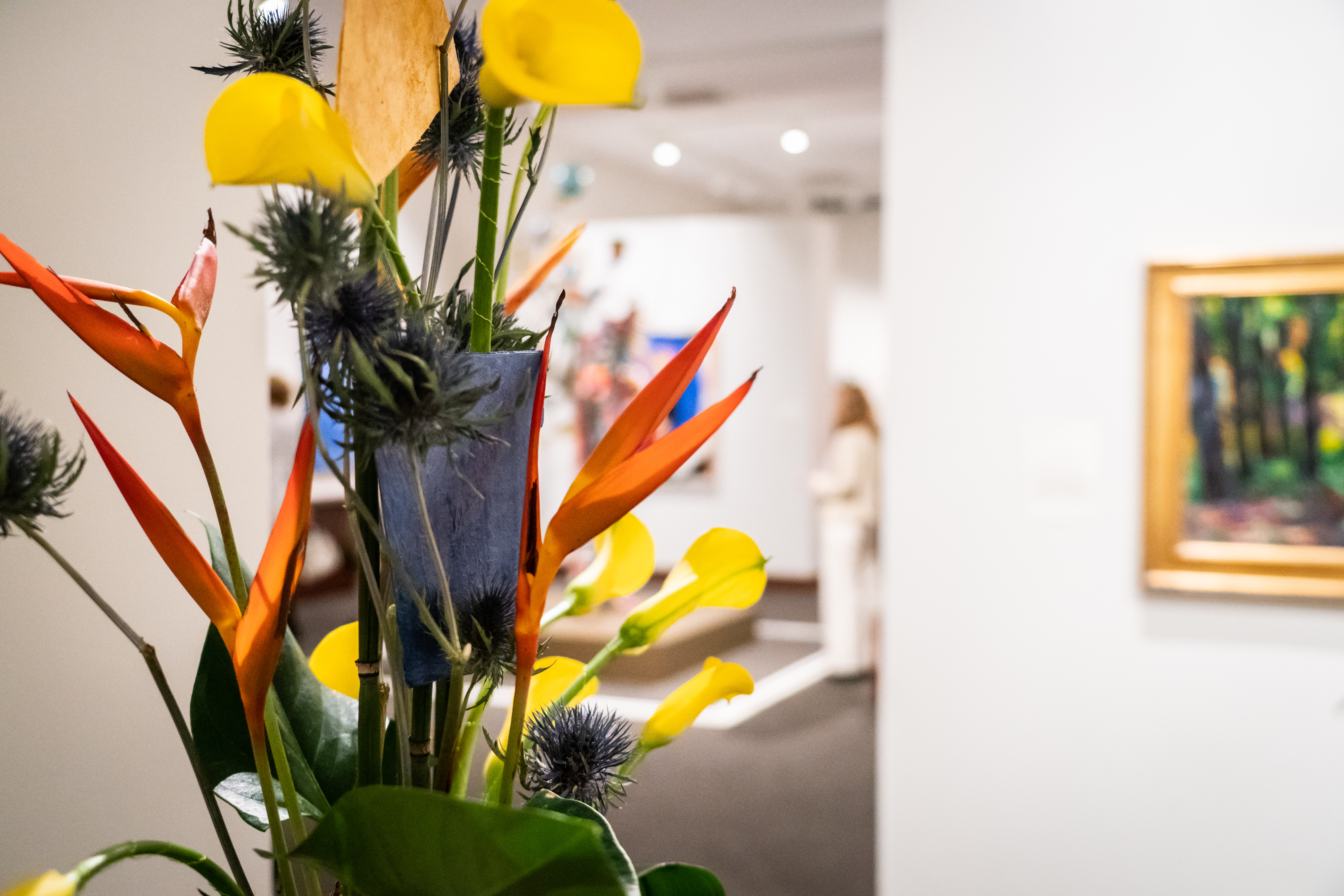 The camera is focused on a colorful floral arrangement in the MAM galleries. Further back and out of focus, a painting is hanging on the wall and a woman is looking at Nick Cave's sound suit.