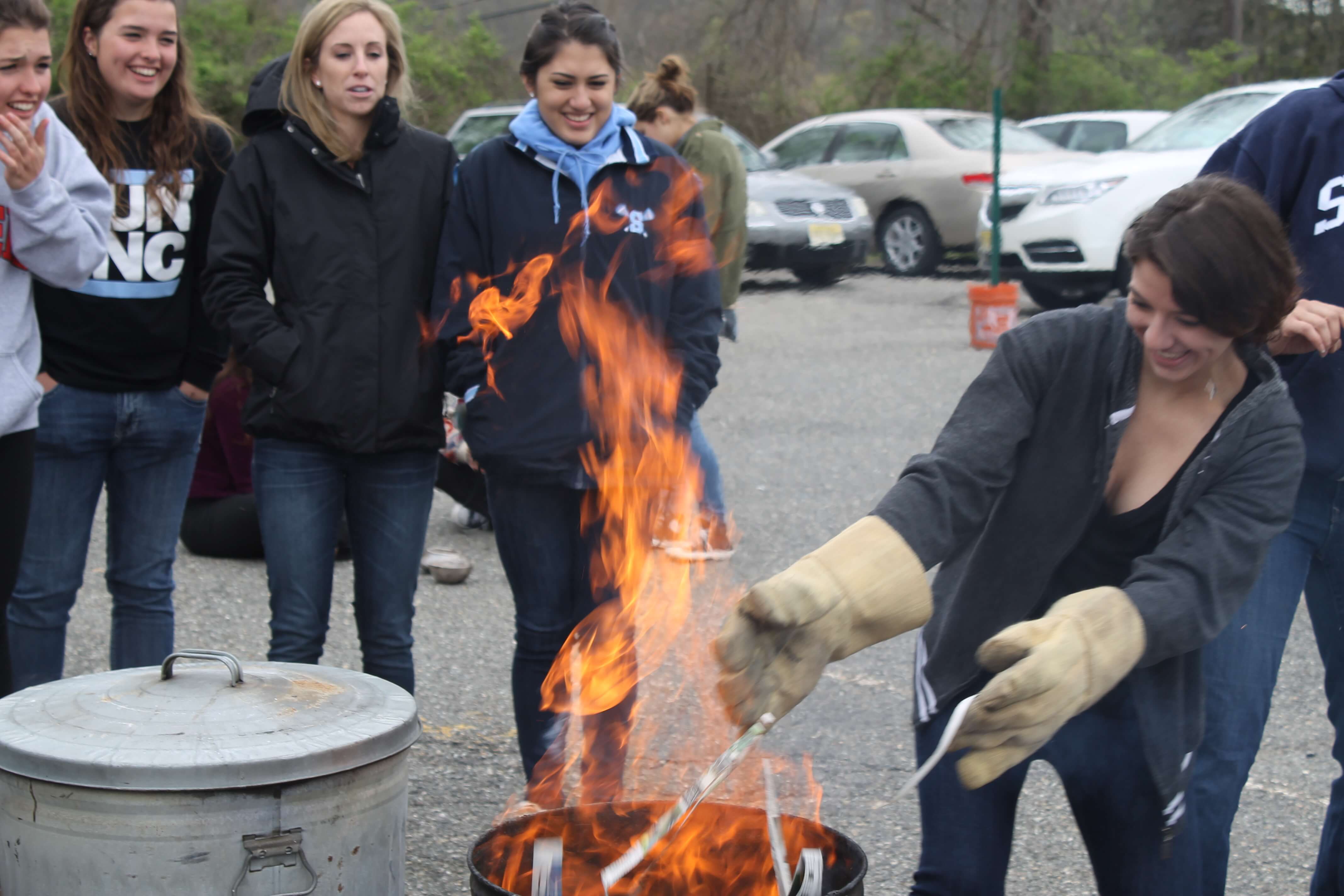Four high school girls are watching as another girl lowers her raku clay piece into a trash can full of burning newspaper. Everyone is wearing winter coats and smiling. 