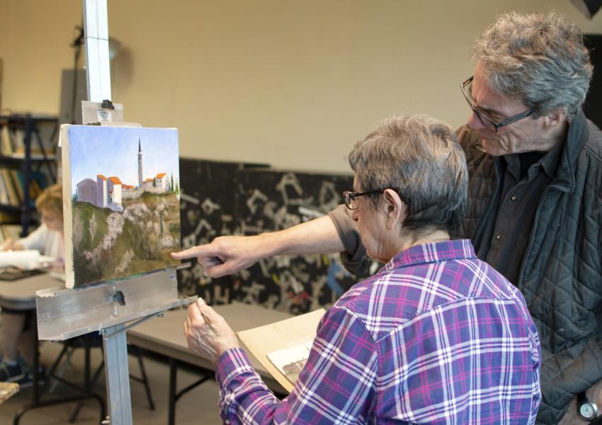 A woman is painting a landscape. Aninstructor is pointing something out to her on the canvas. Both are facing away from the canvas.