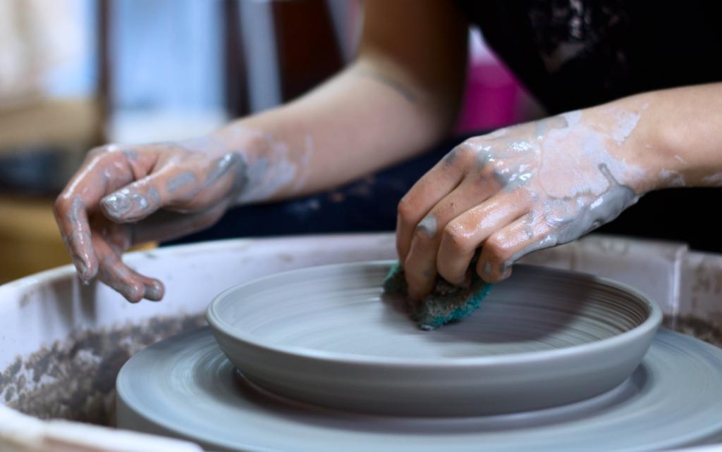 A student is shaping a plate with a sponge on the pottery wheel. Only their hands are visible.