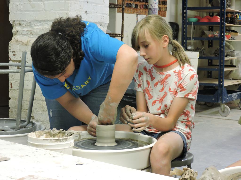 an instructor is showing a student how to make her piece taller on the pottery wheel. The student is watching. 