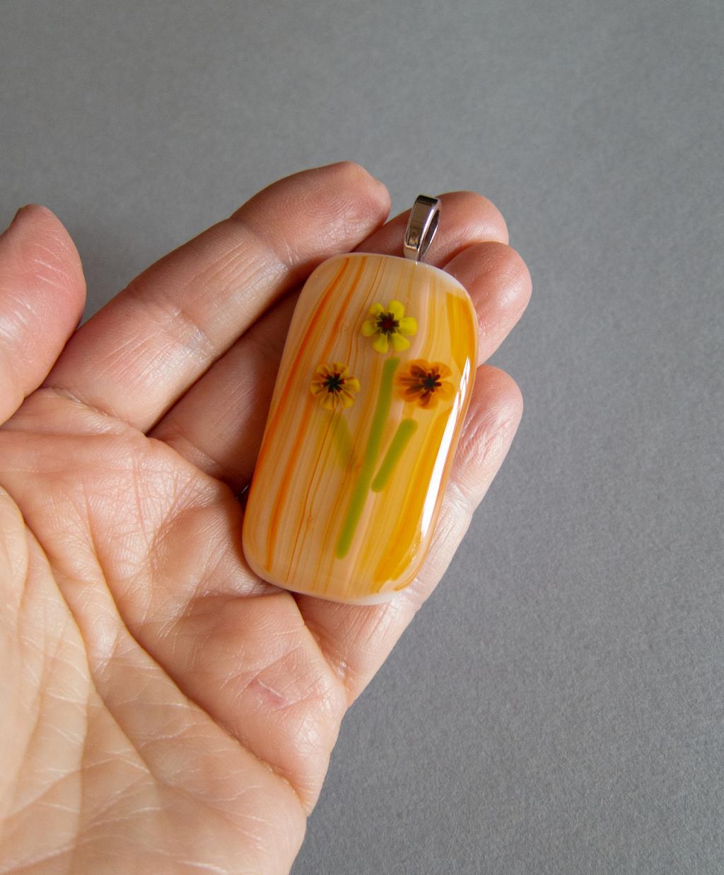 a close up picture of a hand holding a pendant made of fused glass. glass is in a floral pattern with an orange tonal striped background. 