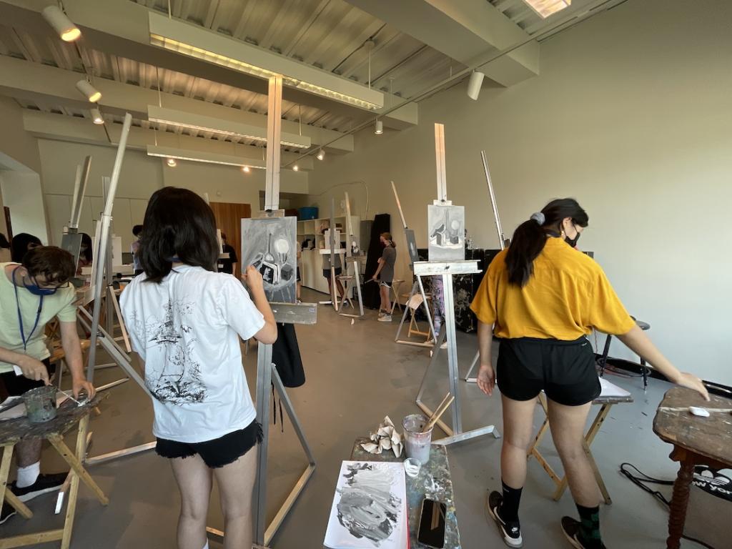 Two students with their backs toward the camera working on still life paintings on easels.
