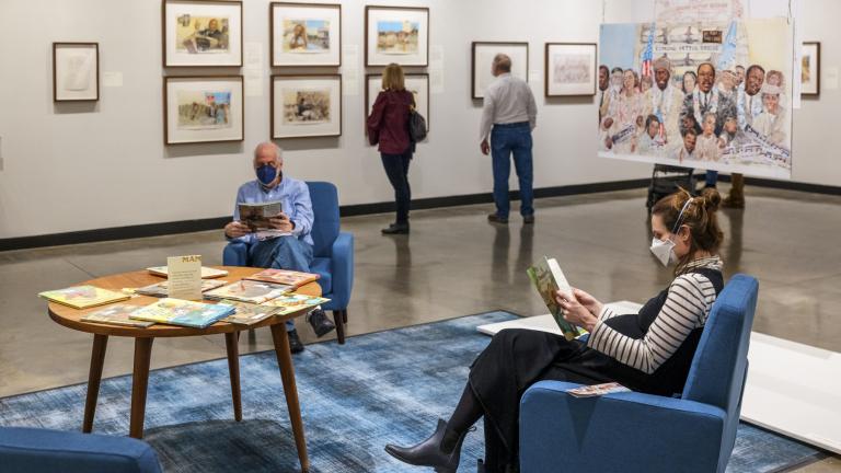 Two visitors reading in chairs in the foreground and more visitors looking at artwork on the wall in the background.