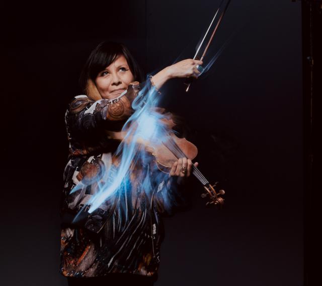 Artful picture of Laura Ortman playing violin against a black background with streaks of light trailing her bow.