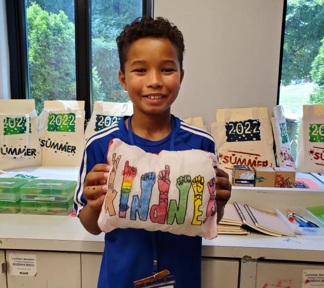 Camper holding up a pillow with a printed "KINDNESS" pressed into it that he designed in SummerART Studio Art Camp