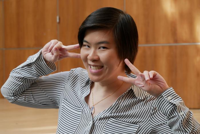 A woman with short black hair is throwing up peace signs on either side of her face and smiling while standing in front of the stage at montclair art museum.