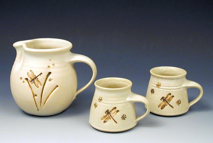 A creme colored pitcher and two mugs, created by hand on the pottery wheel. They've been set up on a backdrop for professional photography. 
