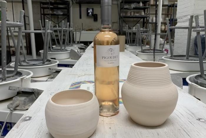 a wine bottle and two handmade pieces of pottery sit in the center table between the rows of pottery wheels in the ceramics studio at MAM.
