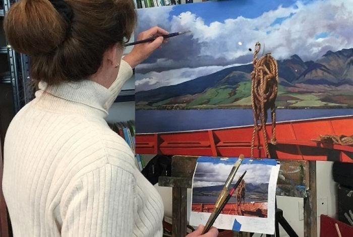 A student is facing away from the camera, painting a lake and mountain scene on an easel.