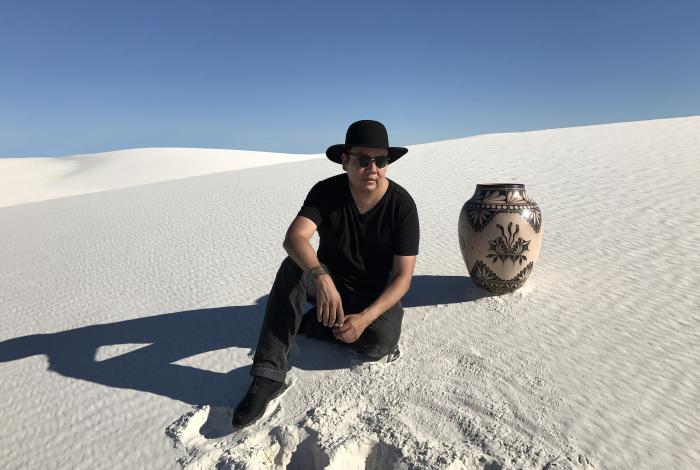 Virgil ortiz is sitting on a sand dune next to one of his elaborately decorated pots that is 1.5-2 feet tall.