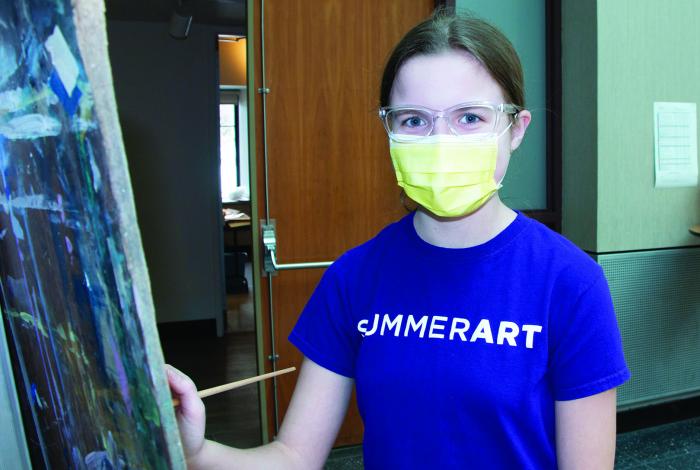 a teen student is painting on an easel and looking at the camera.