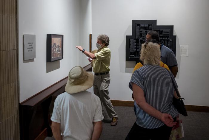A docent pointing to a painting on the wall during a tour. People looking at the painting.