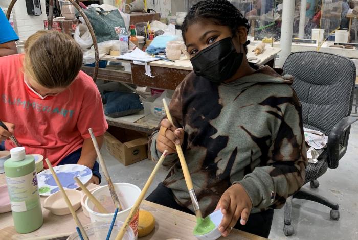 A young student pausing to smile at the camera while glazing a piece of pottery with a brush.