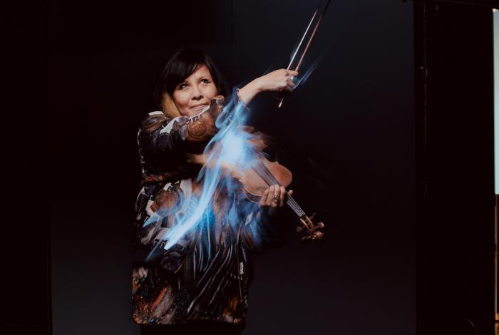 Artful picture of Laura Ortman playing violin against a black background with streaks of light trailing her bow.