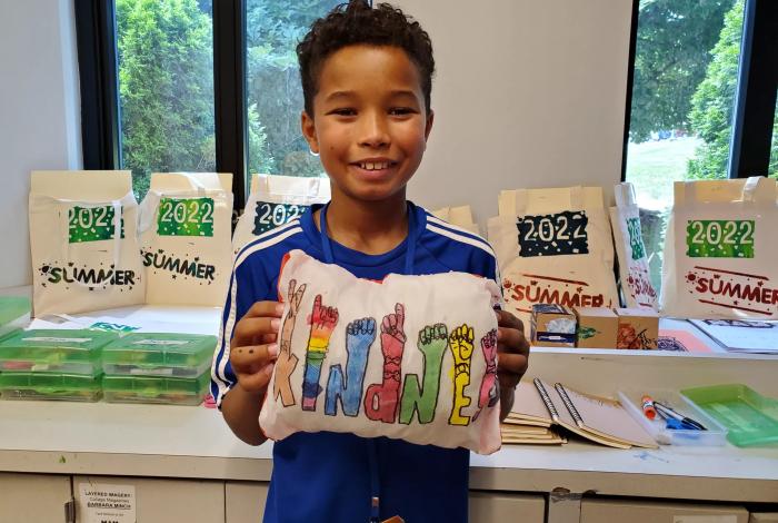 Camper holding up a pillow with a printed "KINDNESS" pressed into it that he designed in SummerART Studio Art Camp